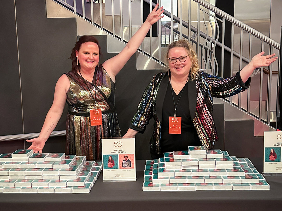 Two women in sequin dresses standing behind a table filled with coasters