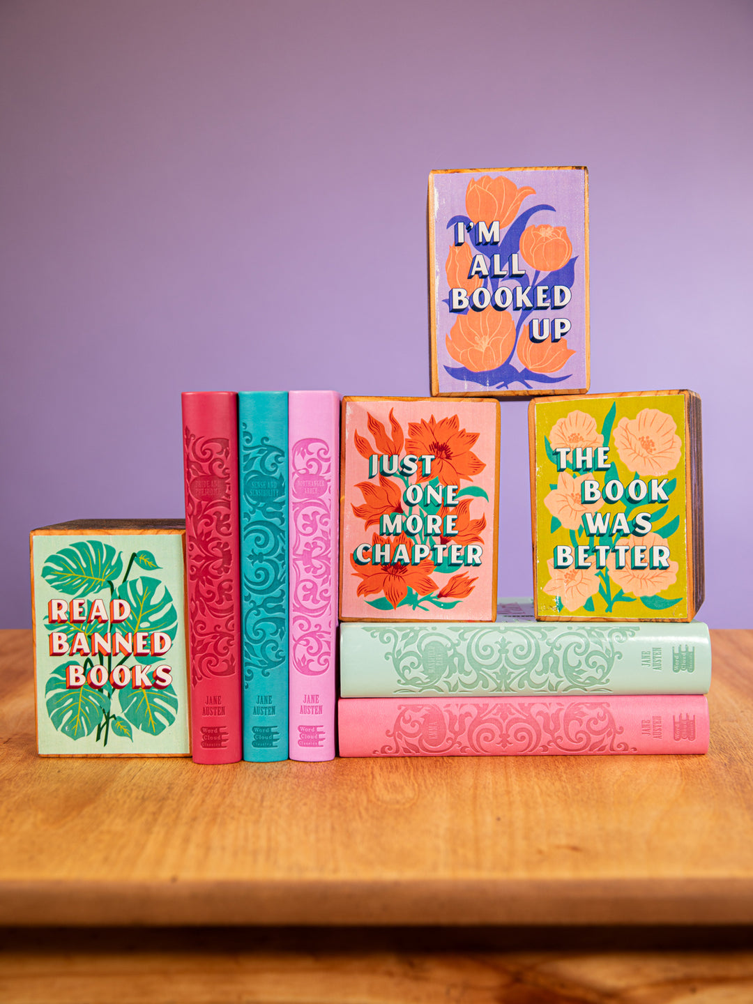 wood table displaying books and wood bookends. Bookends feature bookish quotes "read banned books," "I'm all booked up," "Just one more chapter," and "The book was better." Designs feature bright colors and floral and plant illustrations.