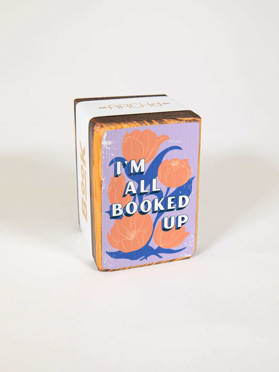 Wood bookend packaged individually in paper sleeve on white background. Bookend face features pink tulip illustration on purple background with bookish quote, "I&