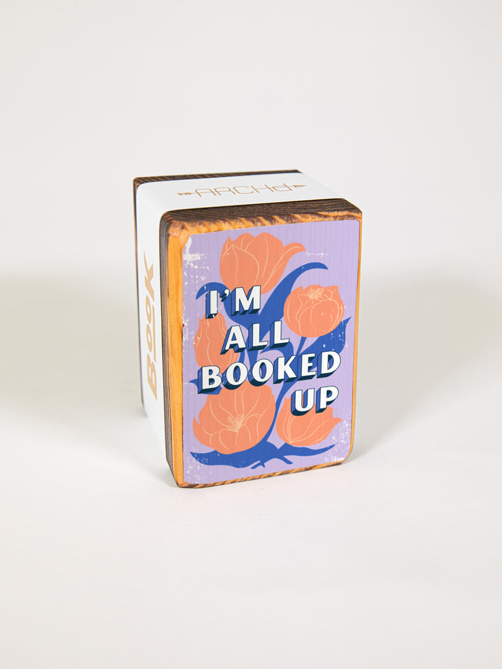 Wood bookend packaged individually in paper sleeve on white background. Bookend face features pink tulip illustration on purple background with bookish quote, "I&