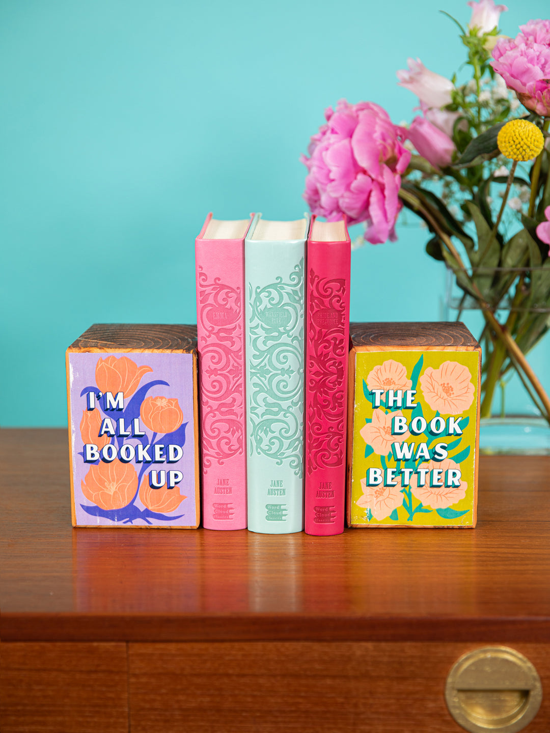 Set of bookends holding up Jane Austen books on coffee table with vase of colorful flowers in background. Bookends feature floral illustrations and book quotes, "I&