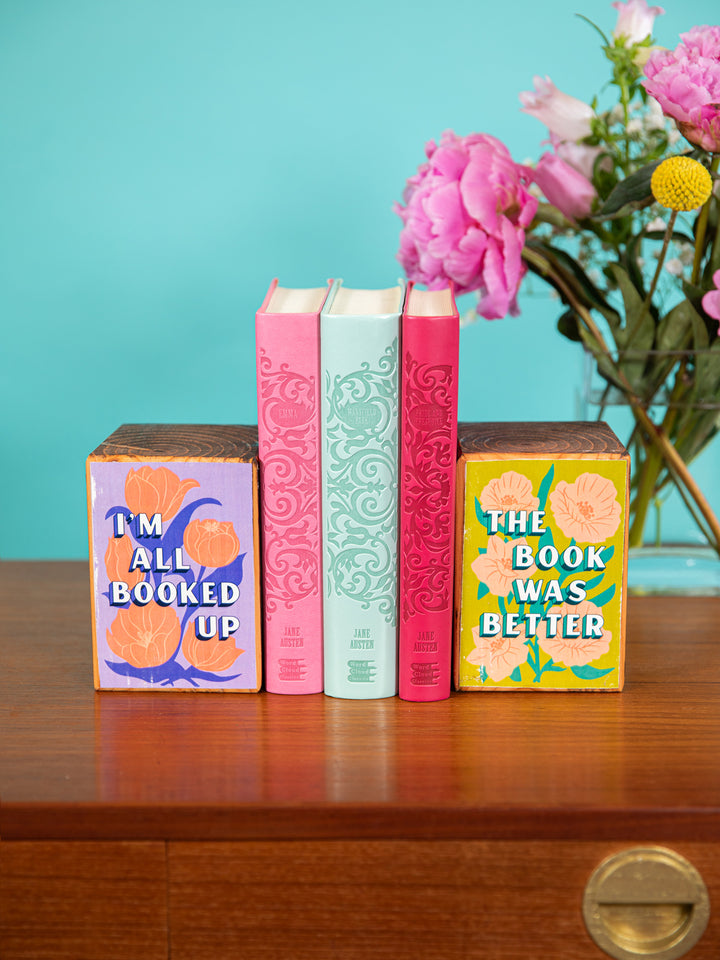 Set of bookends holding up Jane Austen books on coffee table with vase of colorful flowers in background. Bookends feature floral illustrations and book quotes, "I&