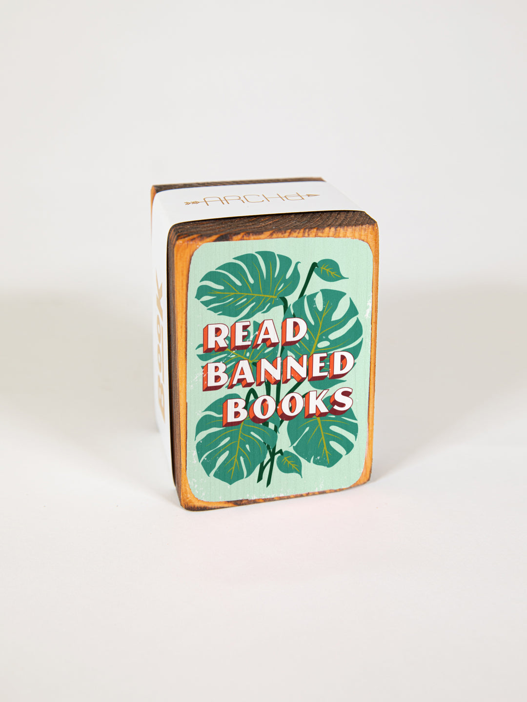 Wood bookend packaged individually in paper sleeve on white background. Bookend face features illustration of green monstera plant leaves with quote, "read banned books," over top of it.