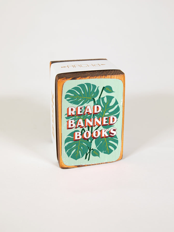Wood bookend packaged individually in paper sleeve on white background. Bookend face features illustration of green monstera plant leaves with quote, "read banned books," over top of it.