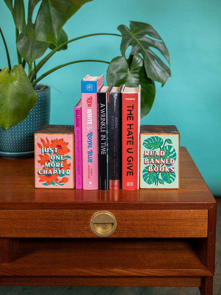 Wood bookends on coffee table with monstera plant in background. Bookends holding up collection of banned books. Bookends feature bookish sayings, "Just one more chapter," and "Read banned books," over floral and plant illustrations.