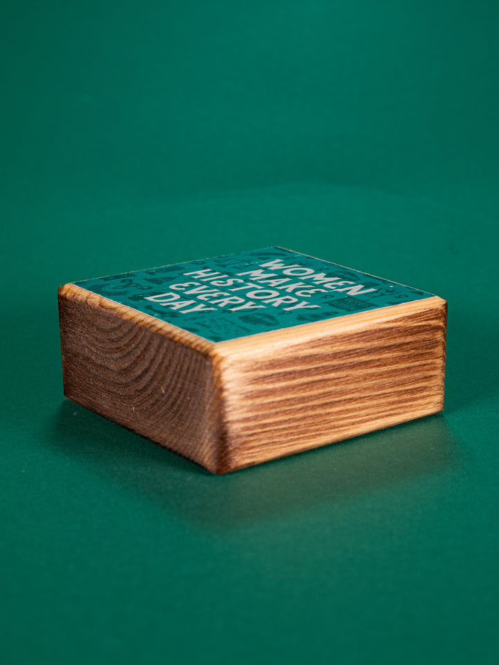 Bottom view of a teal wood block that says "Women Make History Every Day" on a green background