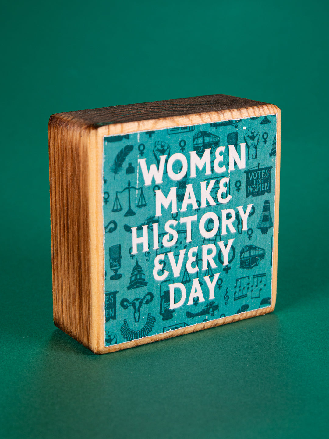 Teal wood block that says "Women Make History Every Day" on a green background