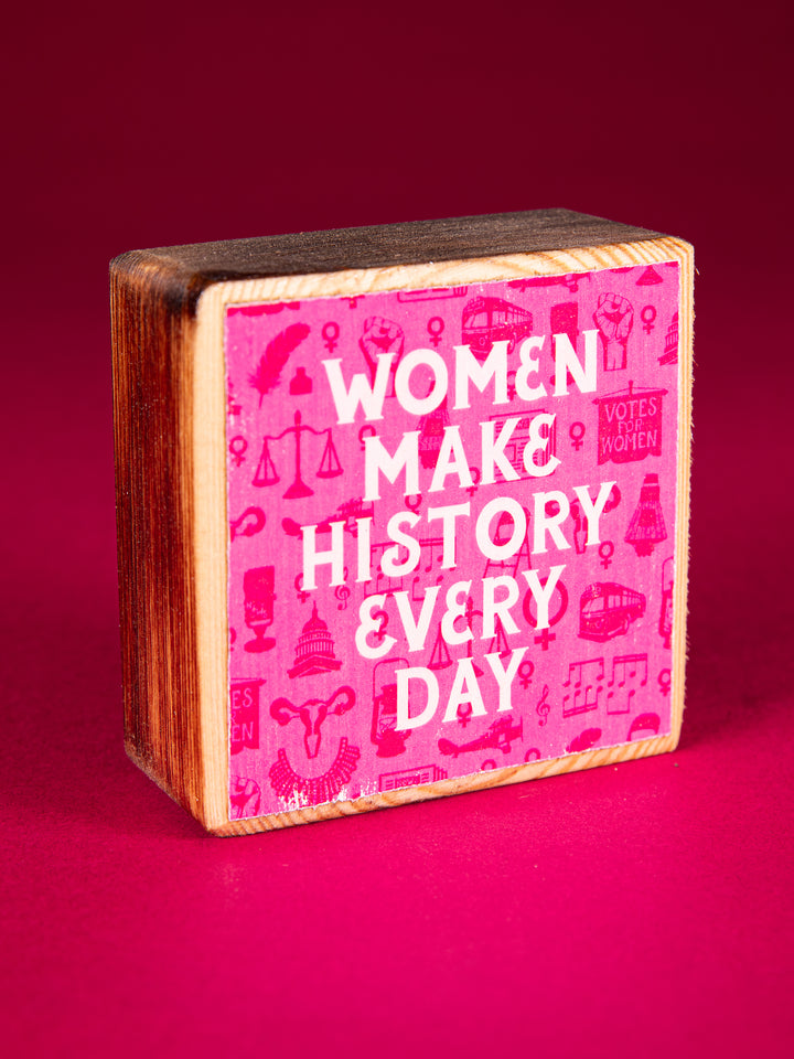 Pink wood block that says "Women Make History Every Day" on a dark pink background