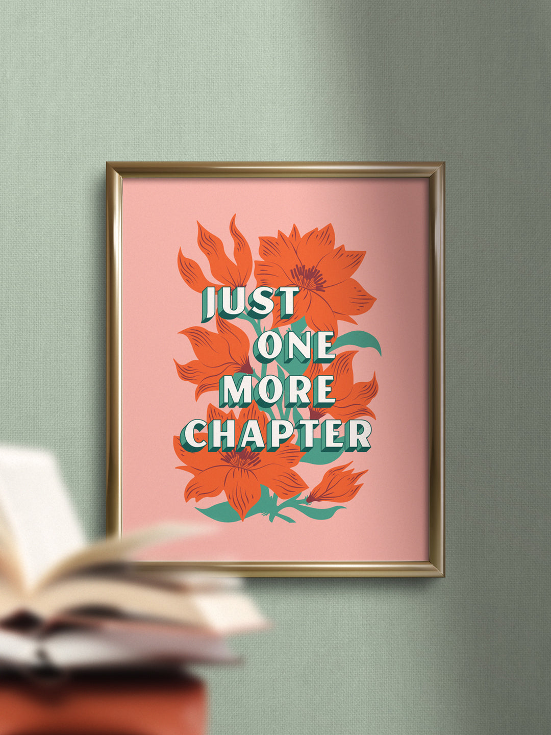 Lifestyle photo of framed art print in gold frame on green wall. Print features bookish saying, "Just one more chapter," over orange wildflower illustration on pink background. Blurred out stack of books in foreground of image.