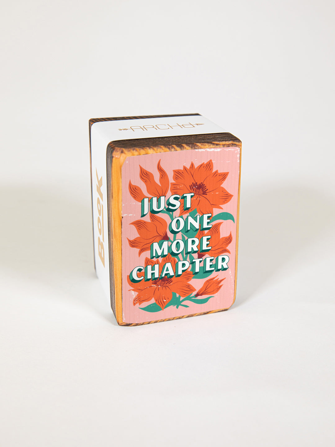 Wood bookend packaged individually in paper sleeve on white background. Bookend face features original orange wildflower illustration on pink background with book lover saying, "just one more chapter."