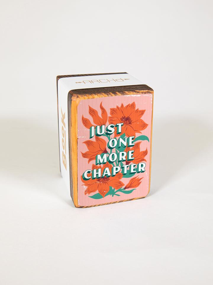Wood bookend packaged individually in paper sleeve on white background. Bookend face features original orange wildflower illustration on pink background with book lover saying, "just one more chapter."