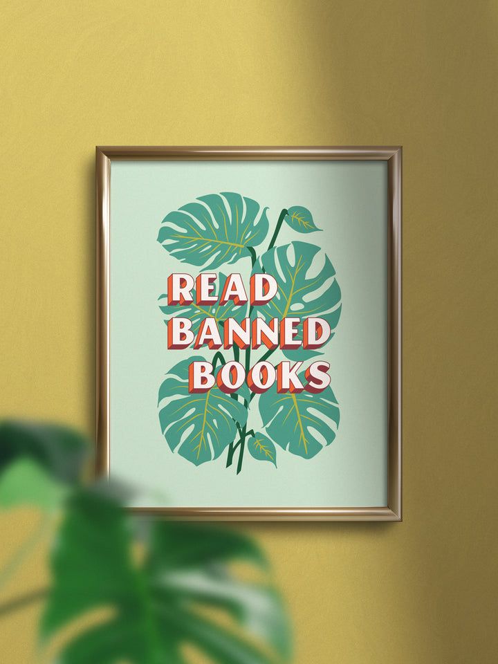 Lifestyle photo shot of art print in gold frame on yellow wall. Art print features bookish quote, "Read banned books," over illustration of monstera plant leaves on mint green background. Blurred out monstera house plant in foreground of image.
