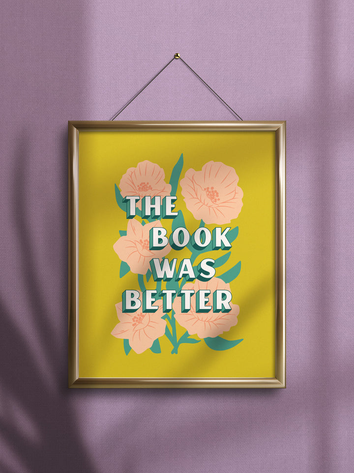 Frame art print featuring yellow/green background with illustration of coral flowers, overlaid with typography of bookish quote, "The book was better." Gold frame hanging from pin on purple wall.