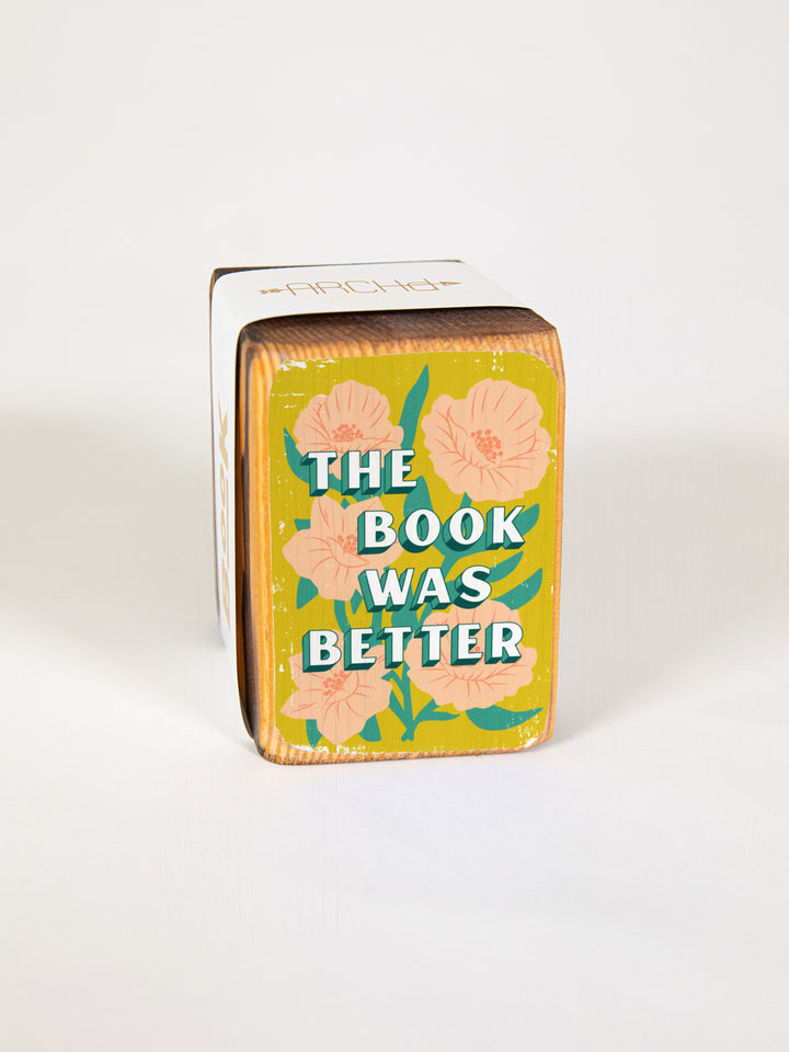 Wood bookend packaged individually in paper sleeve on white background. Bookend face features peony flower illustration with book lover quote, "The book was better."