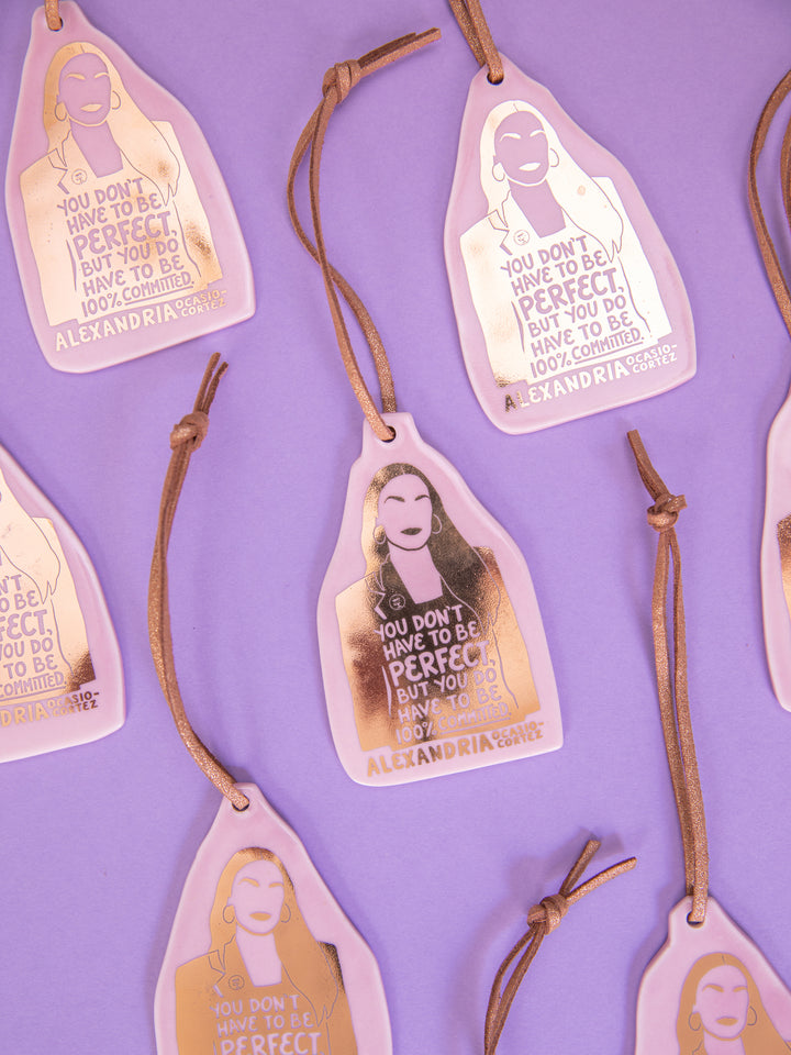 Several Alexandria Ocasio-Cortez ceramic ornaments on purple background. aoc merch