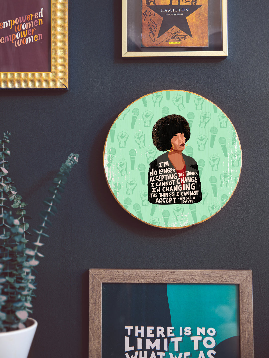 Wood round hanging on black wall with other art surrounding it. Depicting the Angela Davis illustration on a light green background with a microphone and empowerment fist pattern. The quote is "I&