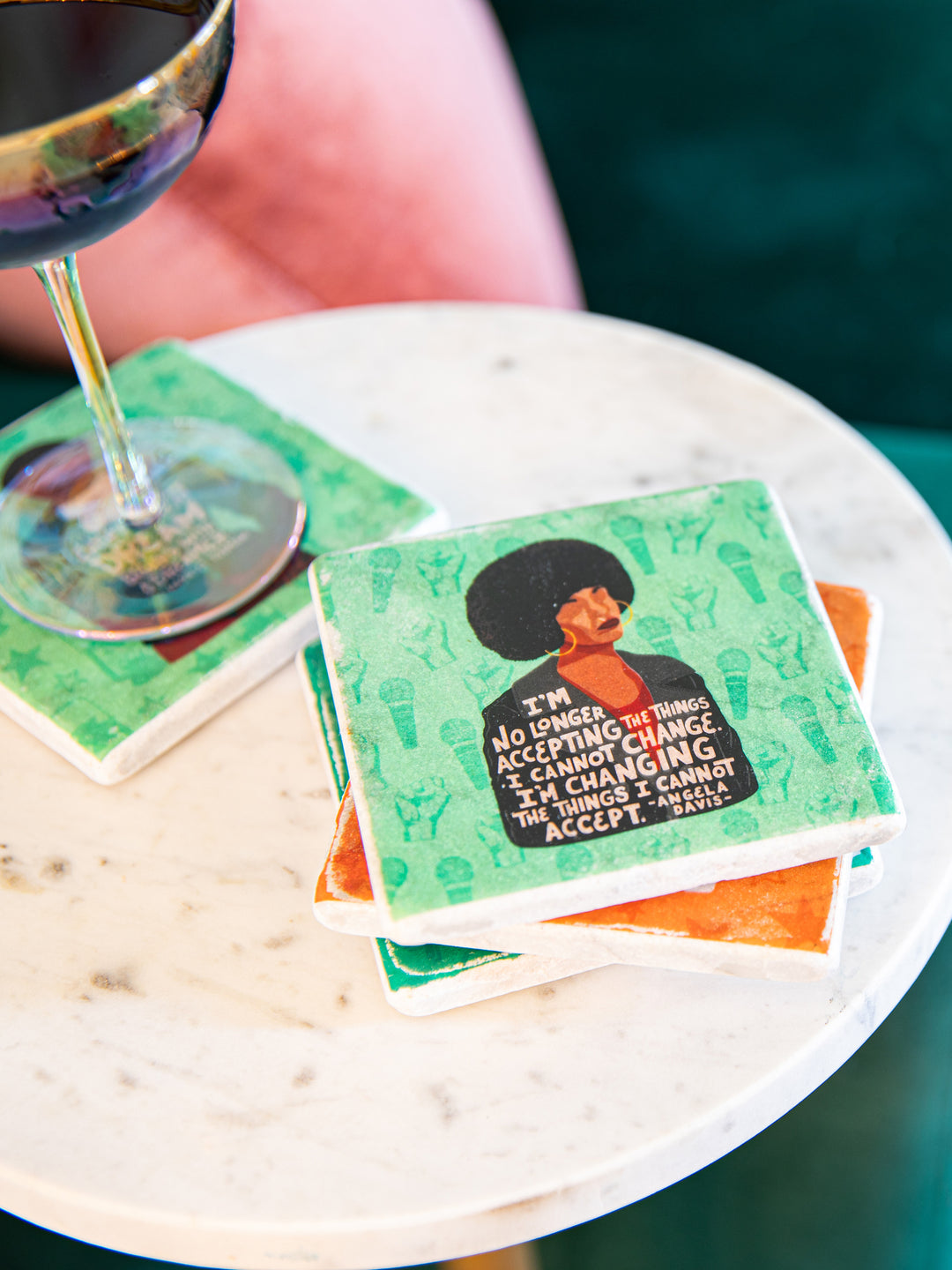 Coasters stacked on a white marble table top. Glass of wine in the background. Front and center is the illustration of Angela Davis on a green background on a marble coaster.