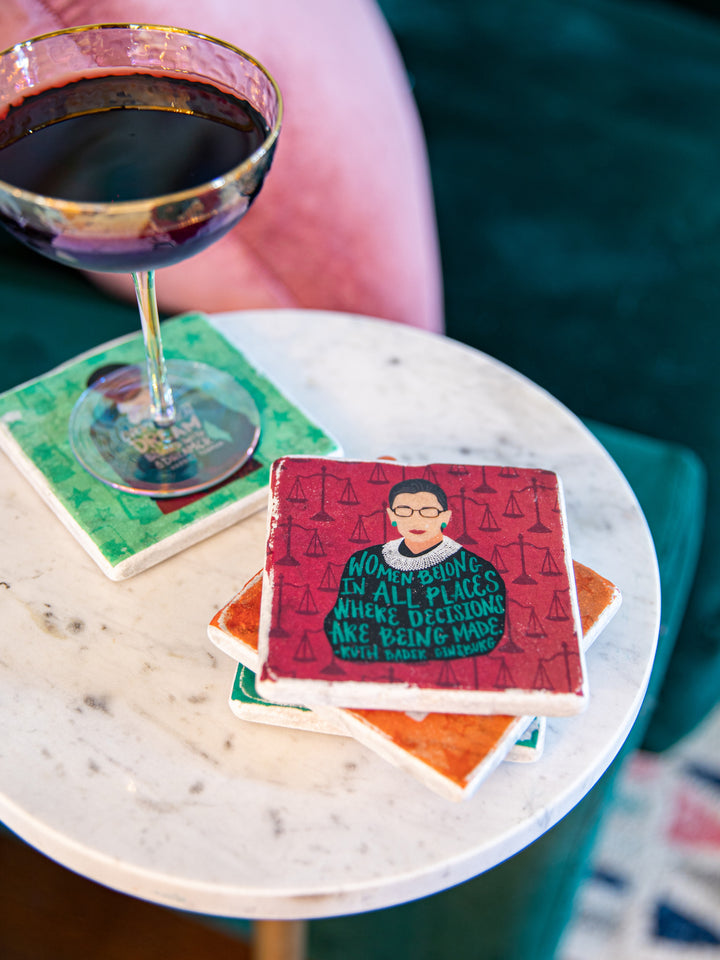 Lifestyle photo of coasters stacked on a marble table top with a glass of wine on one of the coasters. Image shows a RBG coaster front and center on a red background with the scales of justice patterned on the background in dark red.