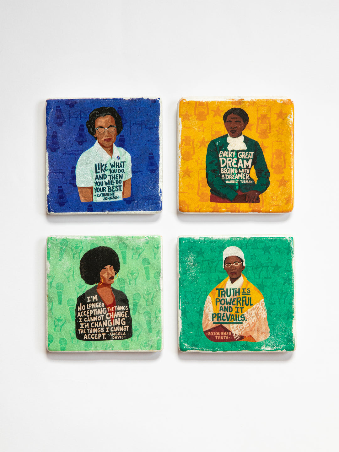 Coaster set of 4 on a white background. Each coaster depicts an illustration of a powerful woman. Katherine Johnson on a blue background, Harriet Tubman on a yellow background, Angela Davis on a light green background, and Sojourner Truth on a dark green background. Each figure has a original quote across their chest.