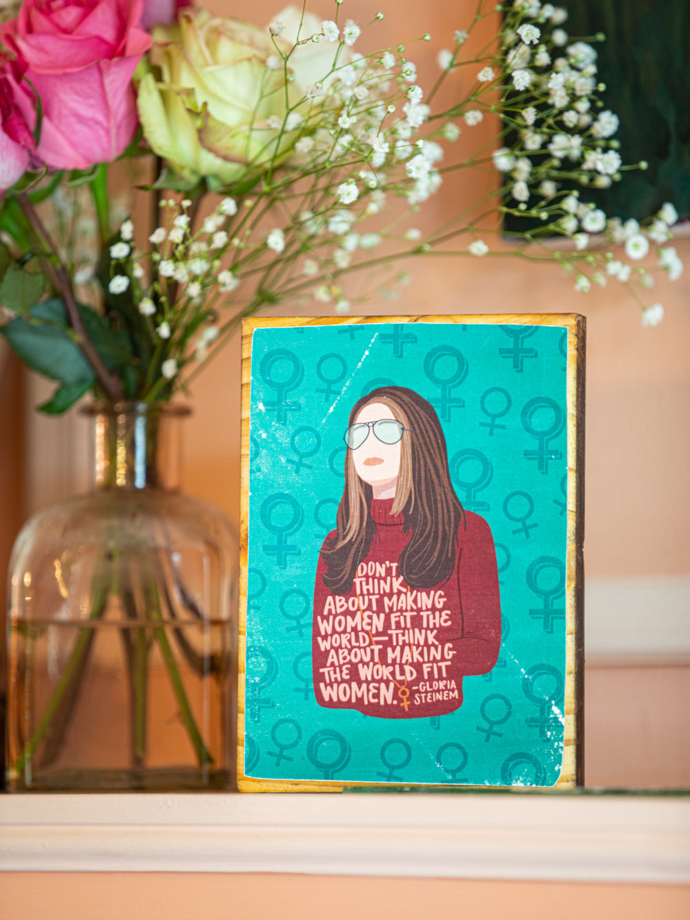 Wood art featuring Gloria Steinem illustration with quote, "Don't think about making women fit the world -- think about making the world fit women." On teal background with illustrations of female symbols. Gloria Steinem art