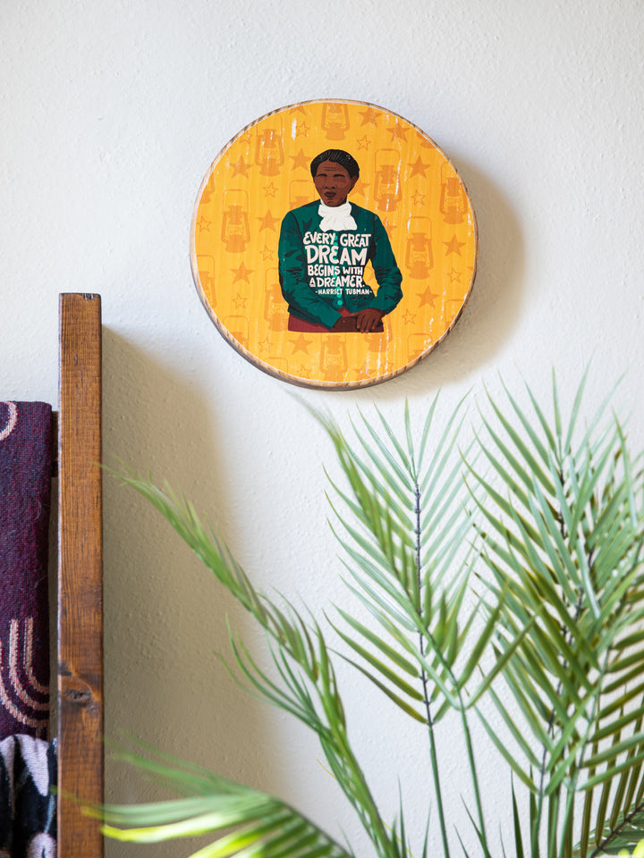 Harriet Tubman wood round illustration on yellow background. Hanging up on a white wall with plants in the foreground.