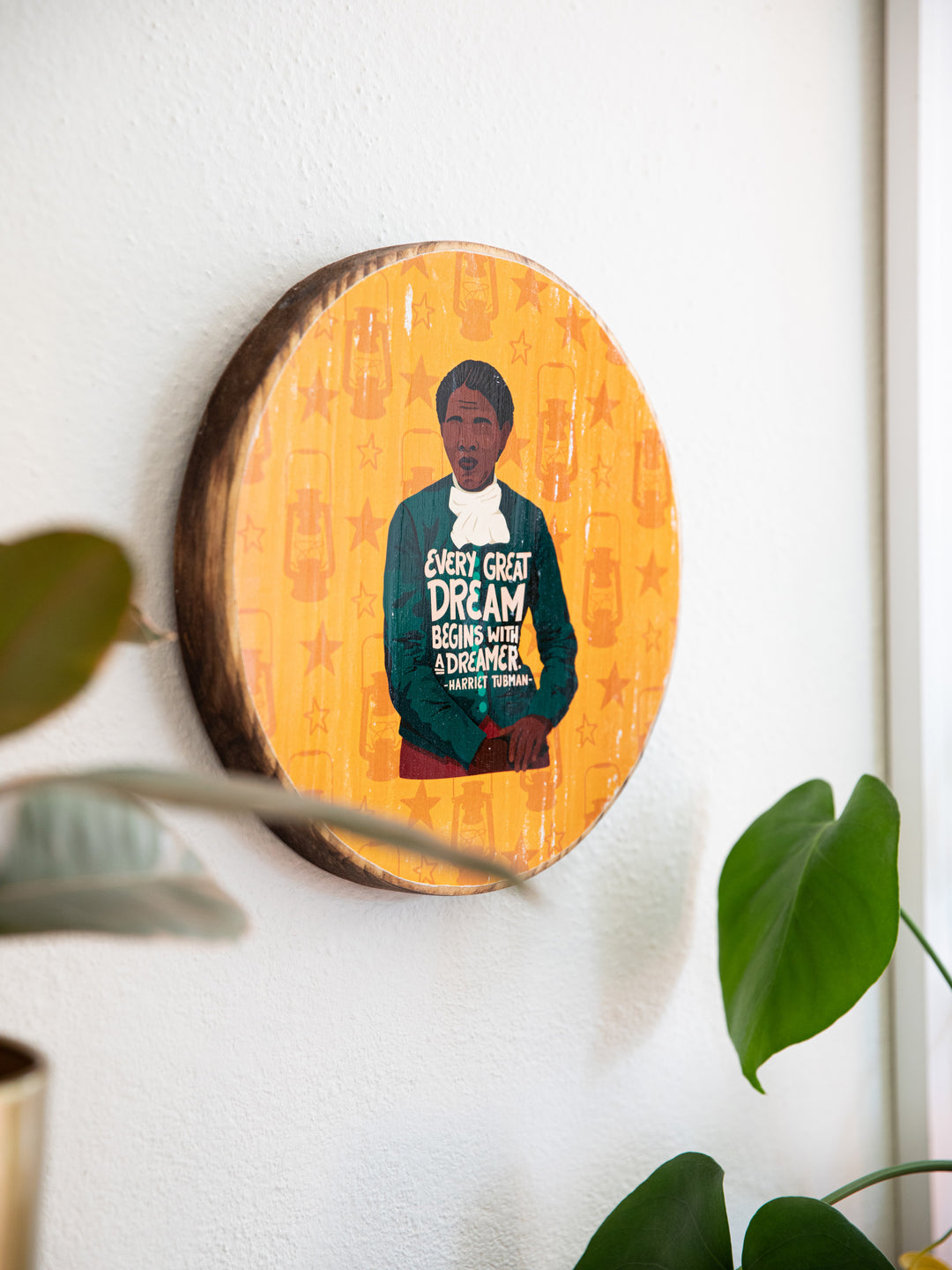 Lifestyle photo. Wood round art hanging on white wall. Depicts Harriet Tubman illustration on yellow background with lantern and star pattern. 