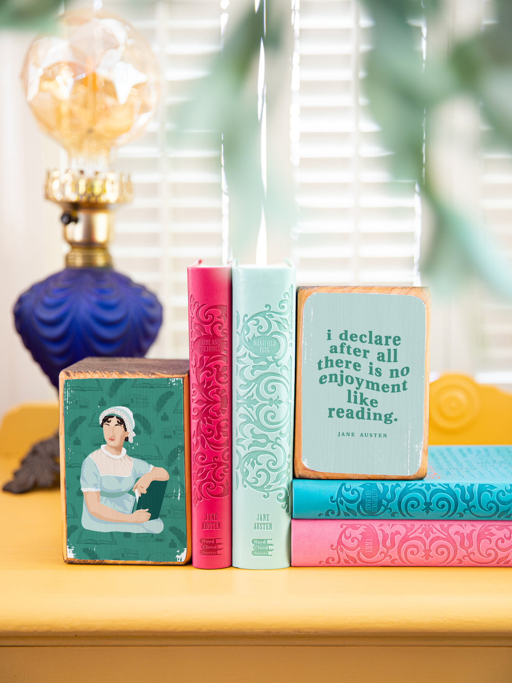 Wood bookends featuring original illustration of English novelist, Jane Austen, paired with Elizabeth Bennet quote from Pride and Prejudice, "I declare after all there is no enjoyment like reading." on a gold side table with plant in the foreground and blue lamp in the background. Jane Austen gifts