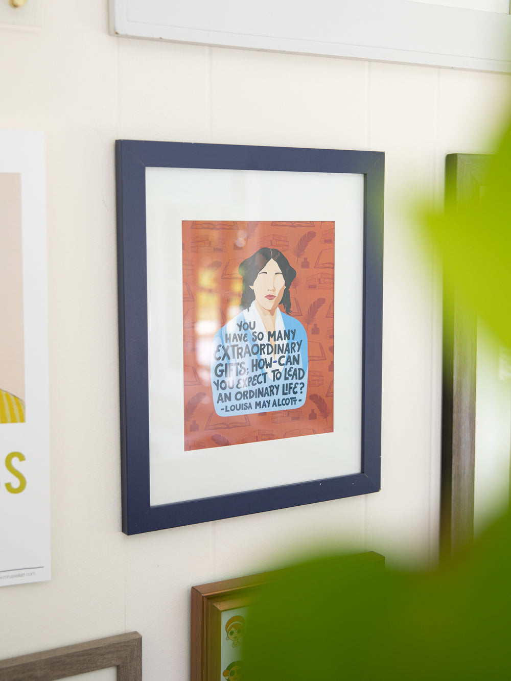 Louisa May Alcott illustrated 8x10 art print on white wall in navy frame with white matte, plant in the foreground. Louisa May Alcott gifts