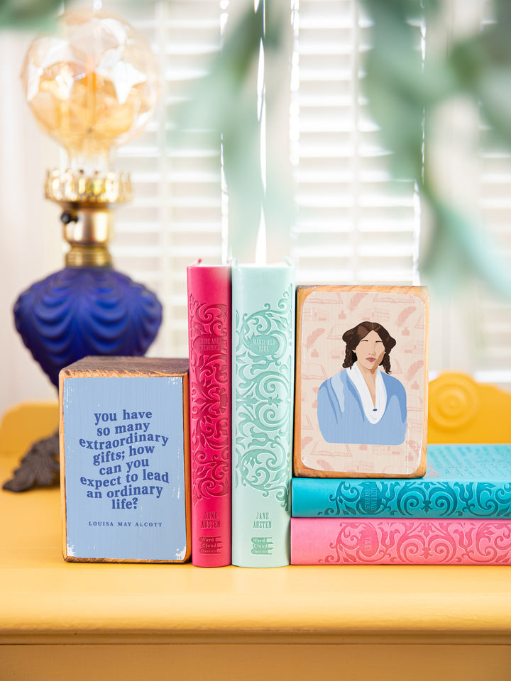 Wood bookends featuring original illustration of American novelist and author of Little Women, Louisa May Alcott, paired with Jo March quote, "You have so many extraordinary gifts; how can you expect to lead an ordinary life?" on a yellow side table holding up multi colored books. Louisa May Alcott gifts