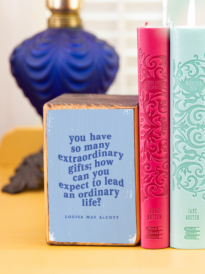 Wood bookends featuring original illustration of American novelist and author of Little Women, Louisa May Alcott, paired with Jo March quote, "You have so many extraordinary gifts; how can you expect to lead an ordinary life?" beside and on top of colorful books, close up of quote bookend. Louisa May Alcott gifts