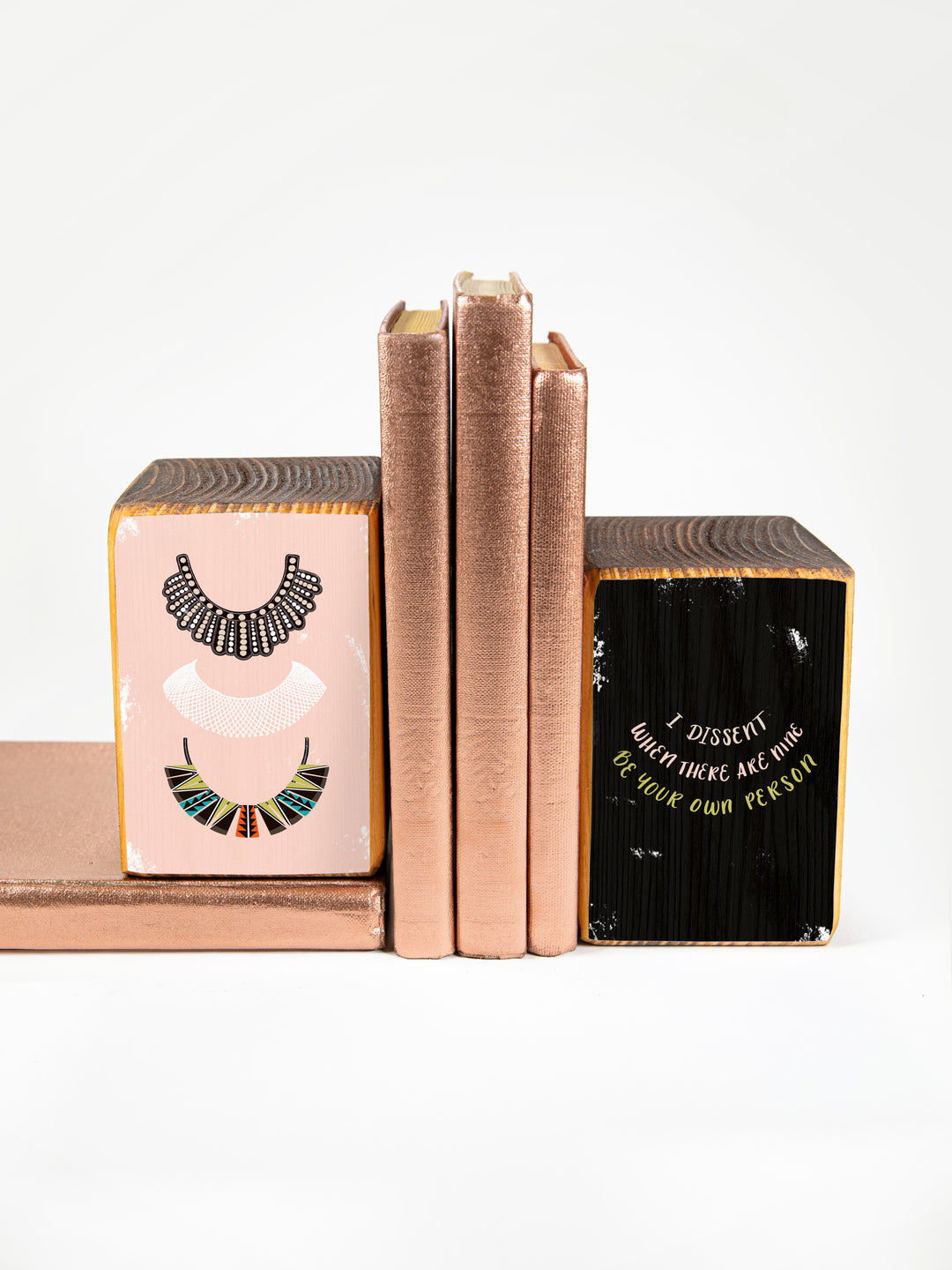 Wood bookend set with illustration of three of Ruth Bader Ginsburg's dissent collars on pink background, paired with RBG sayings, "I dissent," "When there are nine," and "Be your own person," on black background. Bookends displayed on white background, holding up three hardback books. Ruth Bader Ginsburg art