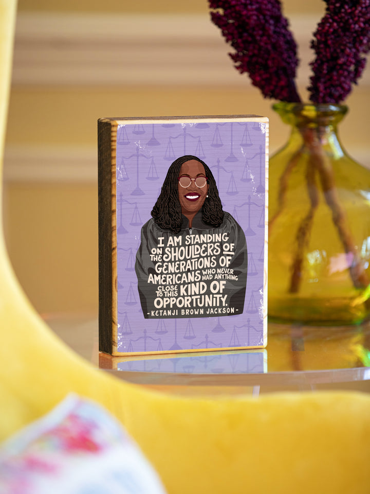 Ketanji Brown Jackson illustration plus quote wood art on a white background on a clear table beside a yellow chair. Ketanji Brown Jackson art