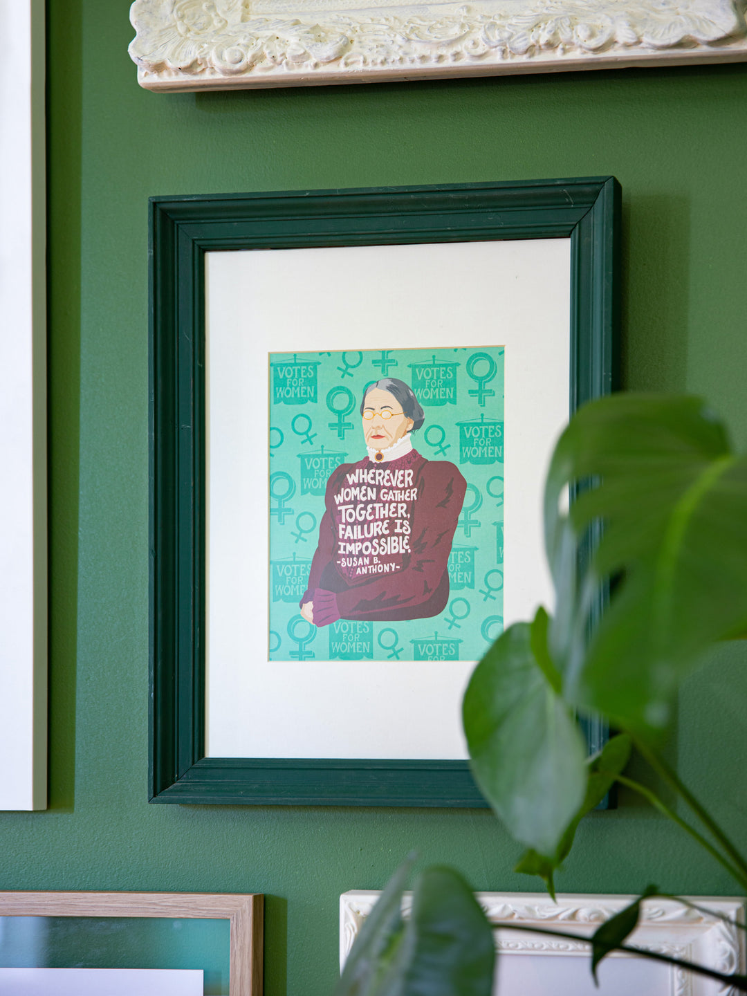 Susan B. Anthony illustrated art print in dark green frame on lighter green wall with plant in the foreground. Susan B Anthony art