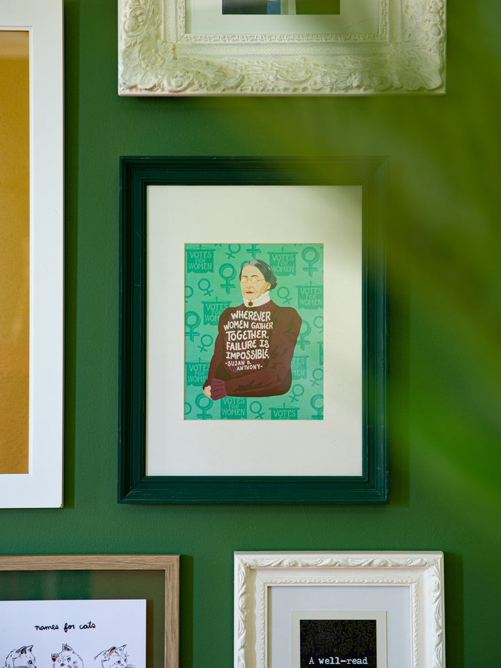Susan B. Anthony illustrated art print in dark green frame as part of a gallery wall with plant in the foreground. Susan B Anthony art