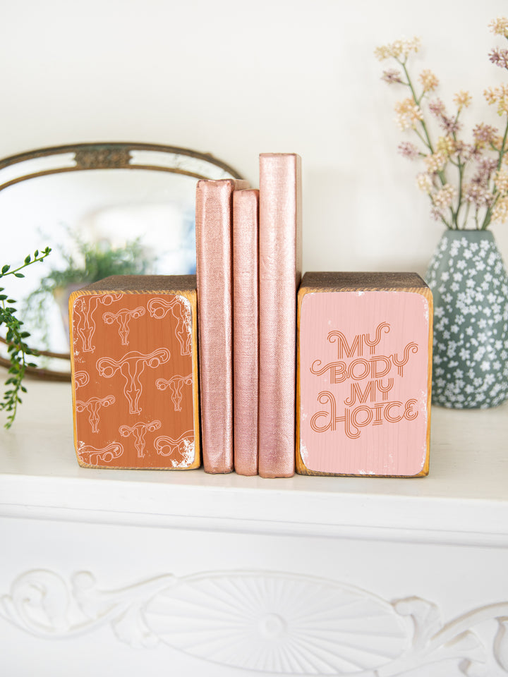 Lifestyle photography of "my body my choice" bookend set on a decorative white mantel, holding up three copper books. wood bookend set of two; left bookend includes repeating pattern of uterus illustrations in pink and copper; right bookend features lettered saying "my body my choice" on light pink background.