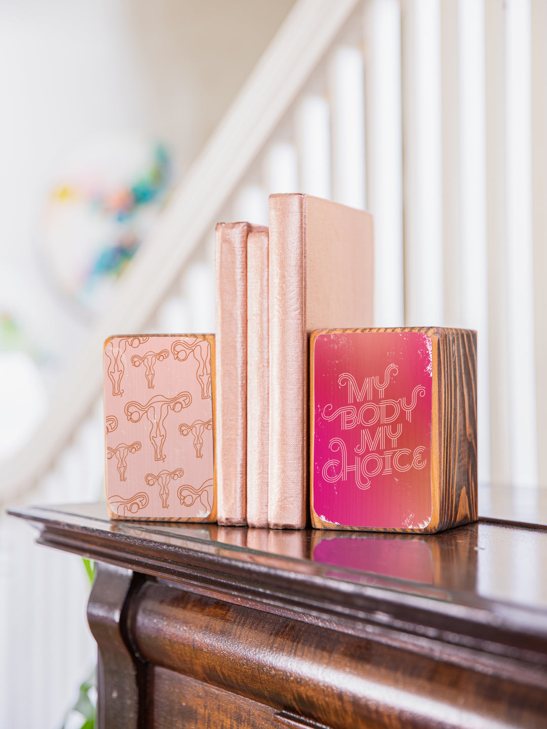 Lifestyle photography of "my body my choice" bookend set on a dark wood piano in front of a white staircase railing, holding up three copper books. wood bookend set of two; left bookend includes repeating pattern of uterus illustrations in light pink and copper; right bookend features lettered saying "my body my choice" on dark pink gradient background.