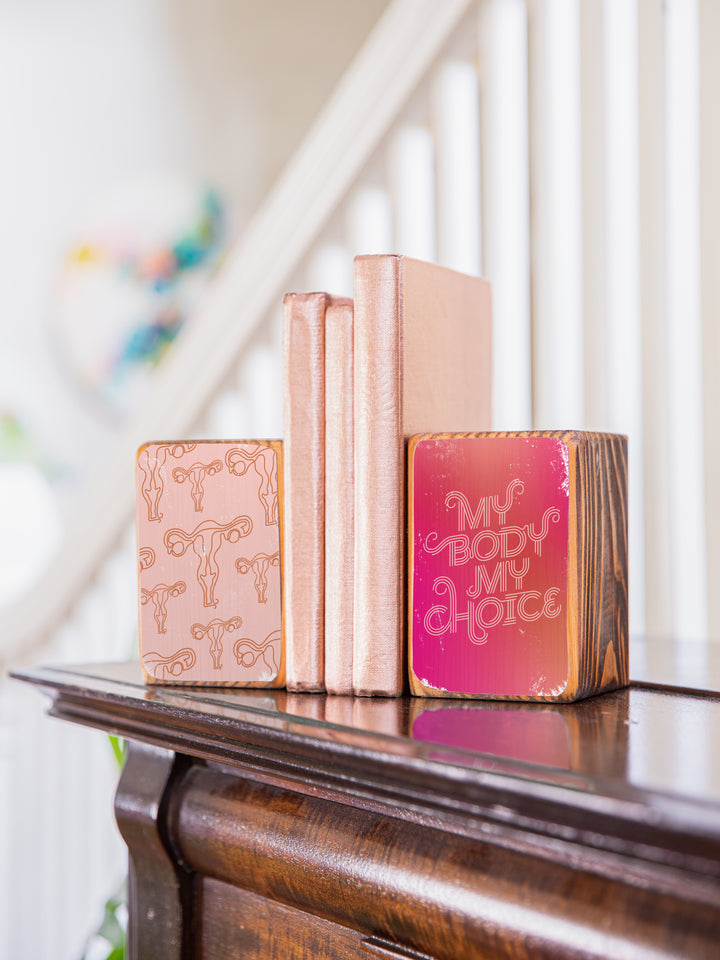 Lifestyle photography of "my body my choice" bookend set on a dark wood piano in front of a white staircase railing, holding up three copper books. wood bookend set of two; left bookend includes repeating pattern of uterus illustrations in light pink and copper; right bookend features lettered saying "my body my choice" on dark pink gradient background.