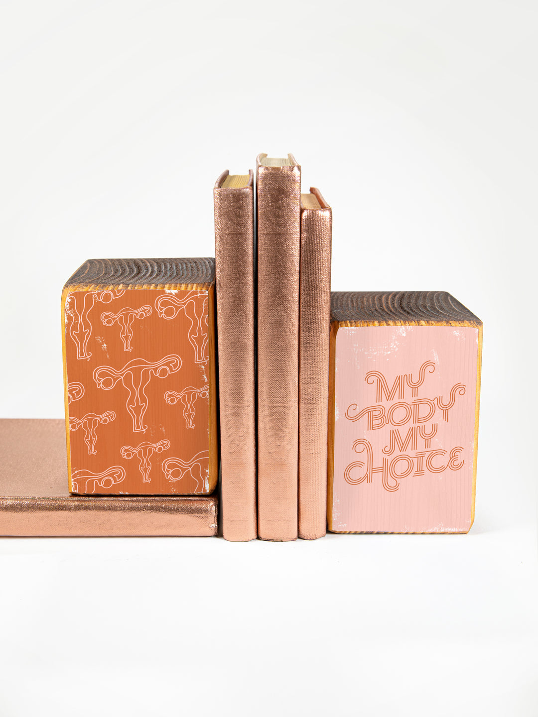 wood bookend set of two; left bookend includes repeating pattern of uterus illustrations in pink and copper; right bookend features lettered saying "my body my choice"; bookend set pictured holding up three copper books against a white background.