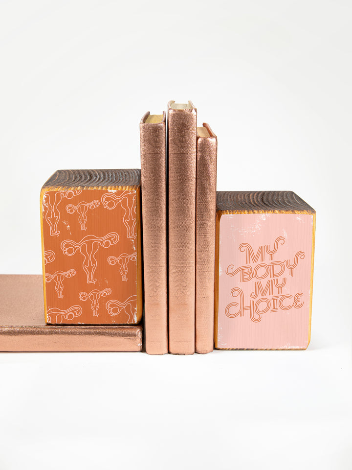 wood bookend set of two; left bookend includes repeating pattern of uterus illustrations in pink and copper; right bookend features lettered saying "my body my choice"; bookend set pictured holding up three copper books against a white background.