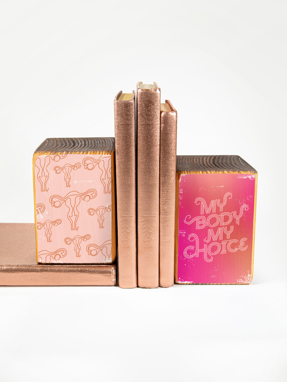 wood bookend set of two; left bookend includes repeating pattern of uterus illustrations in light pink and copper; right bookend features lettered saying "my body my choice" on dark pink gradient background; bookend set pictured holding up three copper books against a white background.